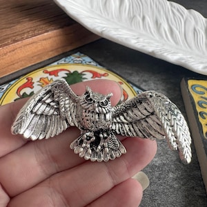 May include: A silver-toned metal brooch shaped like an owl with its wings fully spread. The owl features detailed feather textures on its wings and body, with small, dark, circular eyes and a prominent beak, capturing a flying pose in flight.