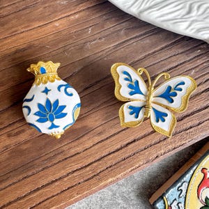 Pode incluir: Dois broches de metal com bordas douradas e detalhes em esmalte branco e azul. Um broche tem o formato de uma bolsa com cordão e motivo floral, e o outro tem o formato de uma borboleta com padrões azuis intrincados nas asas.