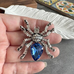 Può includere: Spilla a forma di ragno color argento con cristalli trasparenti e grigi e una grande gemma blu a forma di pera. La spilla è tenuta in mano, mostrando il suo design intricato e i dettagli scintillanti. Un accessorio unico.