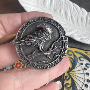 May include: A round, silver-toned metal pendant featuring a detailed raven perched on a branch. The raven is centered within a circular frame with an ornate, swirling border. The pendant has a vintage or antique aesthetic.