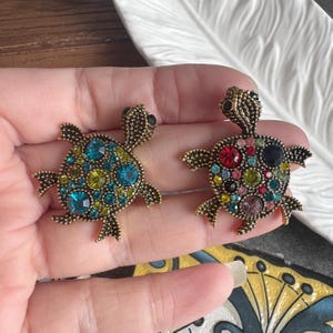 Può includere: Due spille a forma di tartaruga con corpo in metallo color oro antico e superfici testurizzate. La tartaruga a sinistra presenta strass blu e verde lime, mentre quella a destra è decorata con un assortimento multicolore di pietre rosse, rosa, blu e verdi.