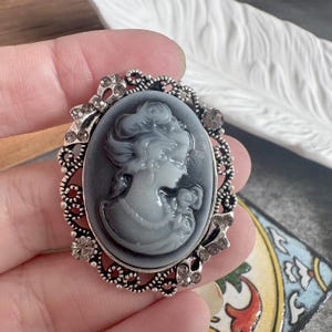 Può includere: Una spilla ovale con un rilievo a cammeo grigio e bianco raffigurante un ritratto di profilo, incastonato in una cornice di metallo color argento. La cornice è decorata con intricati motivi in filigrana traforata e piccoli strass trasparenti.