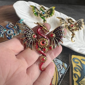May include: A red dragon brooch with metallic gold accents, featuring gear-shaped details, a central red eye, and small red rhinestones on the wings and body. The dragon has spread wings and a curved tail, crafted with a detailed, mechanical-inspired design.