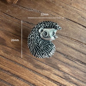 Może przedstawiać: Srebrna broszka w kształcie jeża, o wymiarach 26 mm na 22 mm. Broszka ma szczegółowy wzór z teksturowanym korpusem i uśmiechniętą twarzą. Jeż jest umieszczony na drewnianym tle.
