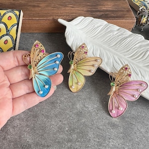 May include: Three butterfly brooches with gold filigree wings and colorful enamel wings in blue, yellow, and pink. Each butterfly has small pearls and red and green accents. The brooches are displayed on a gray surface.