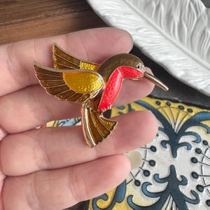 May include: A hummingbird brooch featuring gold-tone metal edges and vibrant enamel fill. The bird has a bright red throat, a yellow chest, and brown wings and tail feathers. The design captures the bird in a mid-flight pose with wings extended.