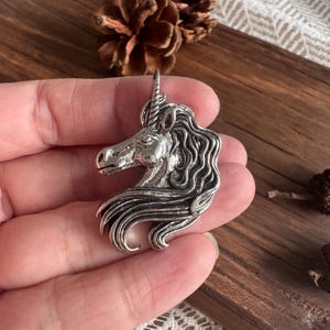 Peut inclure: Une broche en métal argenté en forme de tête de licorne. La pièce présente un profil détaillé de la créature avec une corne en spirale, un œil défini et une crinière texturée et fluide qui s'étend jusqu'à la zone du cou.
