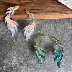 Peut inclure: Deux paires de boucles d'oreilles en forme de poisson avec des contours en métal doré. Une paire présente un émail rose clair et bleu pervenche, tandis que l'autre affiche un dégradé d'émail vert émeraude et bleu sarcelle avec une finition pailletée.