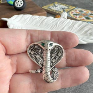 May include: Silver cobra brooch with green and clear crystal detailing. The snake's head is raised, and the body is coiled. The brooch is held in a hand.