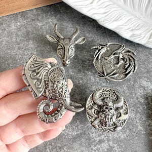 Può includere: Un set di quattro spille in metallo color argento con dettagliati disegni in rilievo. Una spilla raffigura una testa di elefante, una una testa di animale cornuto, una un motivo circolare con drago e una un guerriero vichingo.