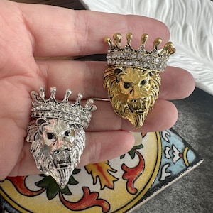 May include: Two lion head brooches, one in a silver tone and one in a gold tone. Each brooch features a detailed lion face with a mane and a crown adorned with clear, sparkling rhinestones. The lion faces have dark accents around the eyes.