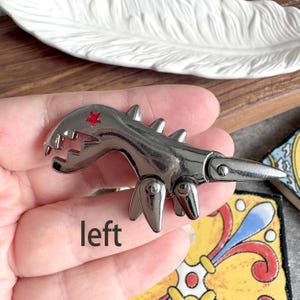 Può includere: Una chiusura per borsa in metallo color canna di fucile a forma di dinosauro con una piccola stella rossa sulla testa. Il fermaglio ha zampe articolate e una coda appuntita, con la parola left stampata in nero sotto l'elemento metallico.