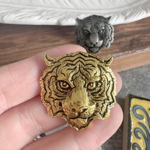 Puede incluir: Un emblema de metal en tono dorado con forma de cabeza de tigre que presenta rasgos faciales detallados, incluyendo patrones de pelaje texturizados, orejas puntiagudas y ojos enfocados. Es una pieza decorativa plana con acabado pulido.
