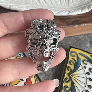 Op de afbeelding: Een zilverkleurige metalen ring met een groot, gedetailleerd schedelontwerp. De schedel heeft een open kaak en is versierd met ingewikkelde, verhoogde filigraanpatronen en kleinere schedelmotieven langs de bovenrand, wat zorgt voor een textuurrijke uitstraling.