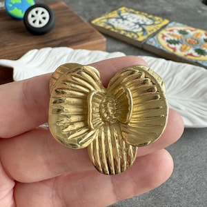 May include: A gold-colored, flower-shaped decorative object. The flower has detailed petal and center designs. The object is held in a hand, with other decorative items in the background, including a wheel and a globe charm.