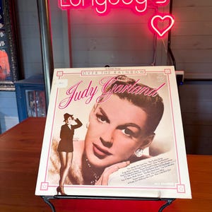 May include: A vintage Judy Garland "Over the Rainbow" album cover on a black metal stand. The album cover features a portrait of Judy Garland and a smaller image of her in a costume. A neon sign that says "Longdog's" is above the album.