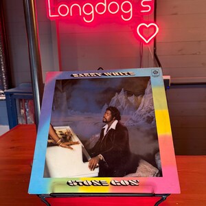 May include: A framed album cover featuring Barry White, with the text "Barry White" and "Stone Gon" visible. The frame has a colorful gradient. A neon sign above reads "Longdog's" with a heart symbol.
