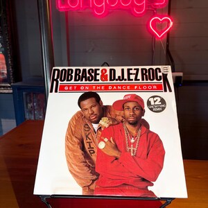 May include: A 12-inch vinyl record cover for Rob Base & D.J. E-Z Rock titled Get on the Dance Floor. The cover features two individuals, one in a brown jacket and one in a red tracksuit with a red cap, wearing gold jewellery and chains.
