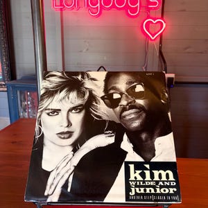 Pode incluir: Uma capa de álbum em preto e branco para "Kim Wilde and Junior" é exibida em um suporte decorativo de metal preto. A capa do álbum apresenta duas pessoas, e o texto "Kim Wilde and Junior" é visível. Uma placa de néon rosa que diz "Longdog's" está no fundo.