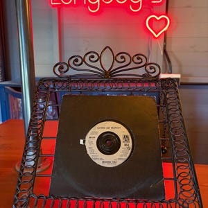 May include: A black vinyl record in a black sleeve is displayed in a decorative wrought iron stand. The record label reads "Chris de Burgh" and "Missing You." A red neon sign above reads "Longdog's" with a heart.