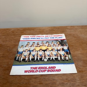 May include: A vintage photograph of the England World Cup Squad. The image shows a team of soccer players in white and blue kits. The text at the top reads "THIS TIME (WE'LL GET IT RIGHT)" and "ENGLAND, WE'LL FLY THE FLAG". The bottom text reads "THE ENGLAND WORLD CUP SQUAD".