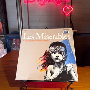 Könnte beinhalten: Ein Schallplattencover für das Original London Cast Album von Les Misérables. Das Cover zeigt eine grafische Illustration einer jungen Person mit Baskenmütze vor einem beigen Hintergrund mit blauen und orangefarbenen Akzenten sowie dem Text A musical that makes history.