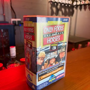 Pode incluir: Caixa de DVD da série britânica "Only Fools and Horses: Série 3". A caixa, amarela, vermelha e azul, mostra o título e fotos dos atores.