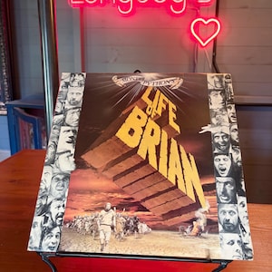 Puede incluir: Una portada de disco de vinilo cuadrada para Monty Python's Life of Brian con grandes letras amarillas en 3D, una escena del desierto con una multitud y un borde de retratos de personajes en blanco y negro. El título dice Monty Python's Life of Brian.