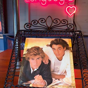 May include: A black metal display stand with a vintage photograph of two men. The stand has an ornate, scrolled design. Above the stand is a neon sign that reads "Longdog's" with a heart symbol.
