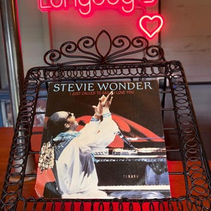 May include: A vintage vinyl record album by Stevie Wonder, titled "I Just Called to Say I Love You," is displayed in a decorative black metal stand. The album cover features a photo of Stevie Wonder performing. A neon sign that says "Longdog's" is in the background.