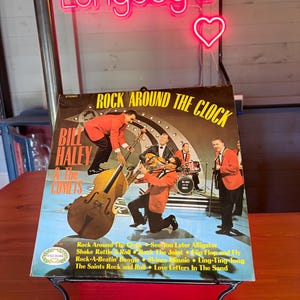 May include: Vintage vinyl record album cover for "Rock Around the Clock" by Bill Haley & The Comets. The album cover features a band in red jackets and black pants. The album is displayed on a black metal stand. A neon sign that says "Longdog's" is in the background.