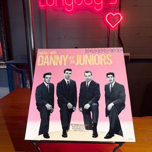 Pode incluir: Uma capa de disco de vinil intitulada Rockin' with Danny and the Juniors, com um fundo rosa e uma fotografia a preto e branco de quatro pessoas com fatos escuros. O texto inclui o nome da banda e a palavra Collectibles.