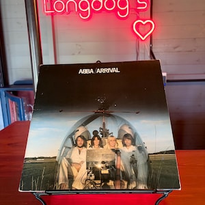 Könnte beinhalten: Ein ABBA Arrival Vinyl-Schallplattencover, das auf einem schwarzen Metallständer ausgestellt ist. Das Albumcover zeigt die Bandmitglieder in einem Hubschrauber. Das Neonschild über dem Album lautet "Longdog's" mit einem Herzsymbol.