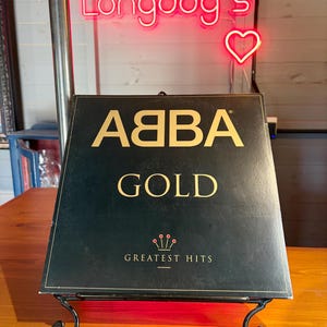 Könnte beinhalten: Ein quadratisches schwarzes Vinyl-Schallplattencover für ABBA Gold Greatest Hits mit goldener Schrift und einem kleinen Kronensymbol. Das Cover steht auf einem schwarzen Metallständer. Darüber leuchtet ein pinker Neon-Schriftzug Longdog's mit Herz.