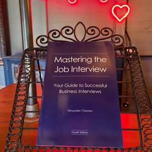 Peut inclure: Un livre bleu foncé intitulé Mastering the Job Interview: Your Guide to Successful Business Interviews par Alexander Chernev, Fourth Edition, posé sur un support en fil métallique noir avec des volutes et un néon rouge au-dessus.