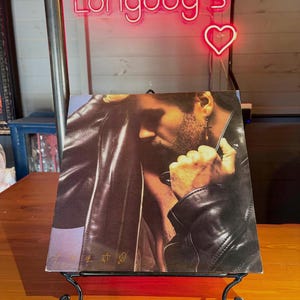 May include: A square vinyl record cover featuring a portrait of a person with a beard wearing a black leather jacket and a cross earring. The cover has a dark, moody aesthetic with gold-colored symbols printed in the bottom left corner.