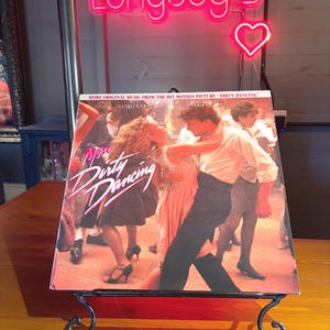 Peut inclure: Une pochette de disque vinyle More Dirty Dancing montrant une scène de danse. Le texte indique More Dirty Dancing et More Original Music From The Hit Motion Picture Dirty Dancing. La pochette est posée sur un support en métal noir.