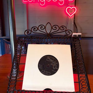 May include: A black vinyl record in a white sleeve is displayed in a decorative black metal stand. The record is labeled "You Are Everything" and is a 45 RPM. A neon sign above reads "Longdog's" with a heart symbol.