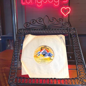 May include: A 45 RPM vinyl record in a white paper sleeve, displayed on a black metal stand. The record label features a mountain graphic, the text Pinnacle, California Dreamin, and Colorado. The label also includes the text 45 RPM and PIN 87.