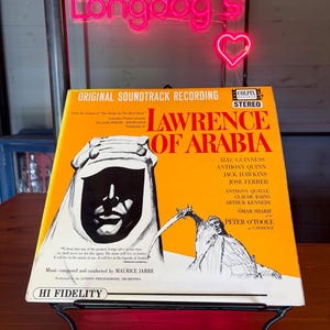 May include: A vintage vinyl record album cover for "Lawrence of Arabia" on display. The album features a striking image of a figure in a headdress and the film's title in bold letters. The album is on a decorative black metal stand.