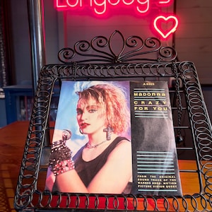 May include: A vintage 7-inch vinyl record sleeve featuring Madonna with blonde hair, wearing a black top and cross earrings. The cover text reads Madonna Crazy For You, A 6323, and From The Original Sound Track Of The Warner Bros. Motion Picture Vision Quest.