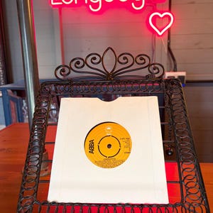 Op de afbeelding: Een vintage ABBA-plaat in een witte hoes, tentoongesteld in een zwarte smeedijzeren standaard. Het label van de plaat is geel met zwarte tekst. Een neonbord erboven leest "Longdog's" met een hartsymbool.