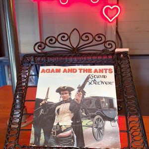 May include: An album cover for "Adam and the Ants Stand & Deliver" in a decorative black metal stand. The cover shows a person in a military-style outfit holding pistols. A neon sign reading "Longdog's" is in the background, with a heart symbol.