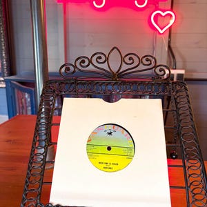 May include: A vinyl record in a white sleeve, displayed in a decorative black metal stand. The record label is yellow and green with the text "WHEN TIME IS STOLEN" and the artist's name, Joan Baez. A neon sign that says "Longdog's" is in the background.