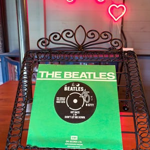 May include: A Beatles 45 RPM record, titled "Get Back" and "Don't Let Me Down," presented in a decorative black metal stand. The record sleeve is green with black and white text. A neon sign in the background reads "Longdog's."