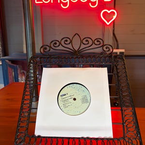 May include: A black wrought iron record display stand holding a vinyl record in a white sleeve. The record has a light blue and white label. A red neon sign above reads "Longdog's" with a heart symbol.