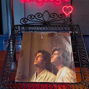 May include: A vintage vinyl record sleeve featuring a portrait of two men in white shirts looking upward, displayed on a black metal wire stand. Above the stand, a bright pink neon sign displays the text "Longdog's" with a heart symbol.