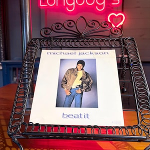 May include: A vintage Michael Jackson Beat It vinyl record sleeve featuring a photograph of the artist in a brown jacket and blue jeans, displayed on a black metal wire stand. The text reads michael jackson and beat it with code A3258.