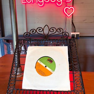 May include: A black wrought iron record stand displaying a vinyl record in a white sleeve. The record label is orange, green, and white. A neon sign above reads "Longdog's" with a heart.