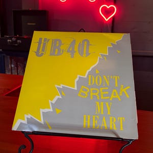 Può includere: Copertina dell'album in vinile "Don't Break My Heart" degli UB40 su un supporto in metallo nero. La copertina dell'album presenta un design giallo e grigio con il nome della band e il titolo della canzone in testo giallo. Un'insegna al neon con la scritta "Longdog's" è sullo sfondo.